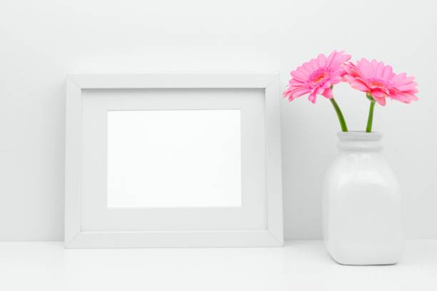 istock Mock up white frame and pink daisy flowers in vase against white wall on shelf 945892646