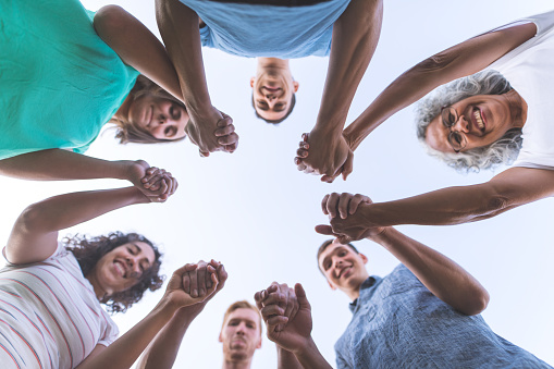 A multigenerational, multiethnic grouping of family put their hands in the middle for a rah rah pump-up cheer. The matriarch, a beautiful older ethnnic woman, is on the upper right side. The shot is from below.