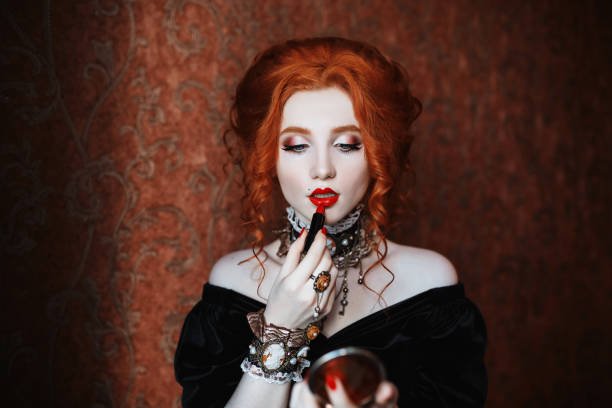 a woman paints lips. vampire with pale skin and red hair in a black dress and a necklace on her neck. girl witch with vampire claws and red lips. gothic look. outfit for halloween. - stage makeup black halloween make up imagens e fotografias de stock