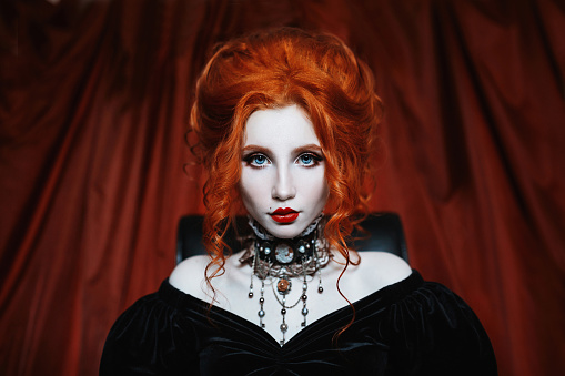 A woman is a vampire with pale skin and red hair in a black dress and a necklace on her neck. Girl witch with vampire claws and red lips. Gothic look. Outfit for halloween.