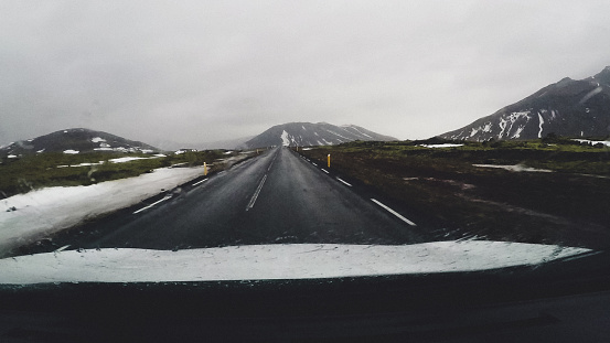 Dashboard camera capturing cold and scenic Iceland. Road is empty and peaceful, and you can really enjoy the time spend here. In distance there are snow-caped mountains, and you can see fog that is coming down on the road. It's raining outside as the wipers are active, but the path is clear and you can see snow on the surrounding meadow and mountains.