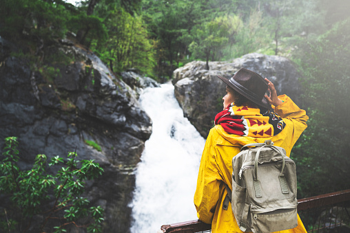Young woman wearing yellow raincoat and backpack hiking in the forest on a rainy day, standing in front of waterfall