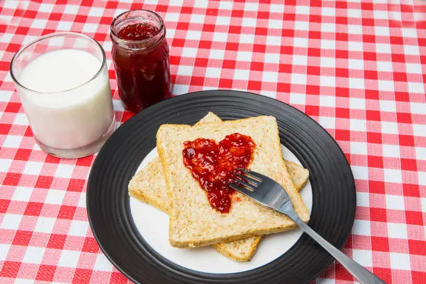 Close up of strawberry jam shaped a heart symbol on a toast bread with milk on the table