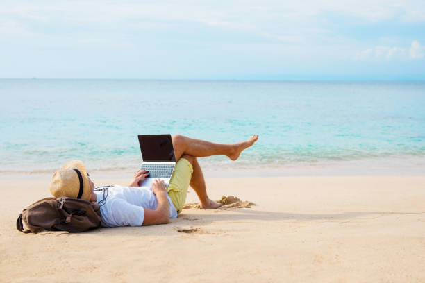 hombre trabajando en ordenador portátil mientras se relaja en la playa - lugar no específico fotografías e imágenes de stock