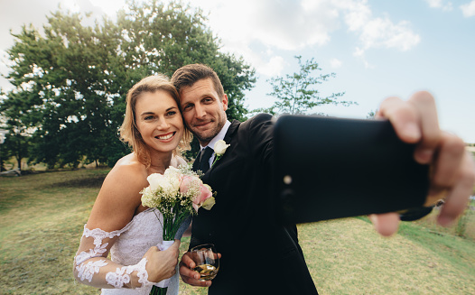 Beautiful bride and groom taking a selfie with mobile phone outdoors. Happy husband and wife do selfie on wedding day.
