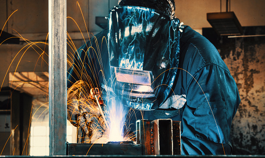 Laborer in protective mask and uniform welds manually metal frame details in shielding gases at production plant close view