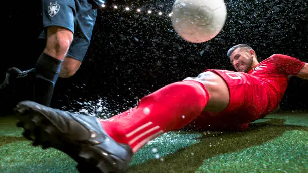 Soccer player trying to slide tackle his opponent in the wet soccer field at night.