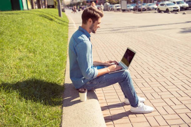 istock happy young man sitting on parapet and chatting on laptop 946047248