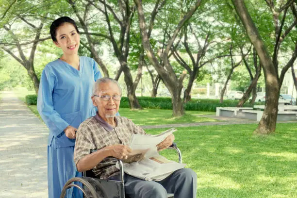 Cheerful female nurse pushing an elderly man on the wheelchair while relaxing in the park