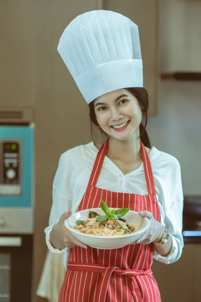 istock Beautiful young woman in chef hat is make Stir-fried spicy spaghetti, looking at camera and smiling while baking in kitchen at home, also known as drunken spaghetti or Phad Kee Mao Spaghetti 946087018