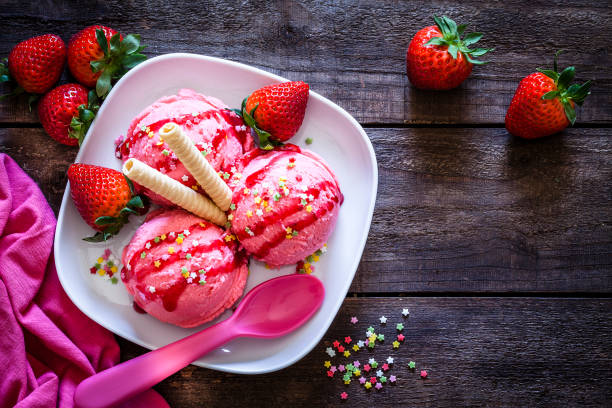 Strawberry ice cream shot on rustic wooden table Top view of three strawberry ice cream balls in a white plate shot on rustic wooden table. The ice cream balls are covered with strawberry syrup.  Some strawberry fruits are scattered on the table around the plate. A pink plastic spoon is on the plate beside the ice cream balls. Natural light used. Predominant colors are pink and brown. DSRL studio photo taken with Canon EOS 5D Mk II and Canon EF 100mm f/2.8L Macro IS USM food fruit close up strawberry stock pictures, royalty-free photos & images