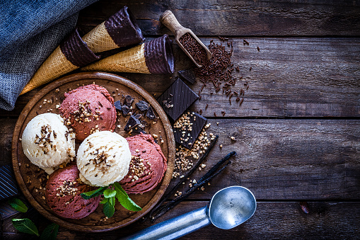 Top view of chocolate and vanilla ice cream balls shot on rustic wooden table. The ice cream balls are covered with chocolate sprinkles and ground hazelnut. Chocolate pieces and chocolate sprinkles are scattered on the table. Three ice cream cookie cones are behind the plate. An ice cream serving scoop complete the composition. Predominant color is brown. DSRL studio photo taken with Canon EOS 5D Mk II and Canon EF 100mm f/2.8L Macro IS USM