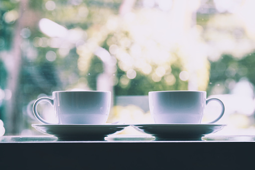 Two White Coffee Cups placing together on table near window with bokeh from outside; picture style film grain