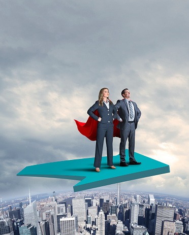 A businesswoman and a businessman each wearing a red cape stand proudly and attentively as they stand with their hands on their hips on a large green arrow that flies above a large urban skyline.