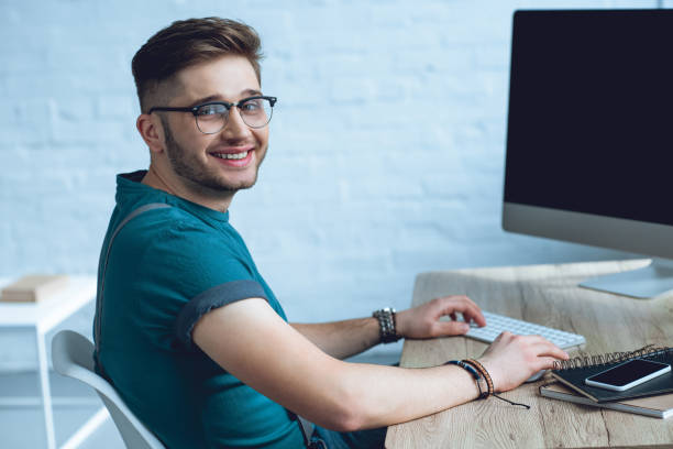 istock handsome young man in eyeglasses working with desktop computer and smiling at camera 946132530