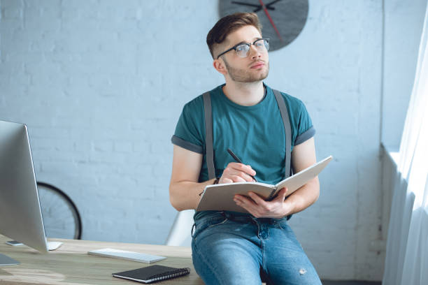 istock pensive young man in eyeglasses looking away while writing in notebook at home office 946137134
