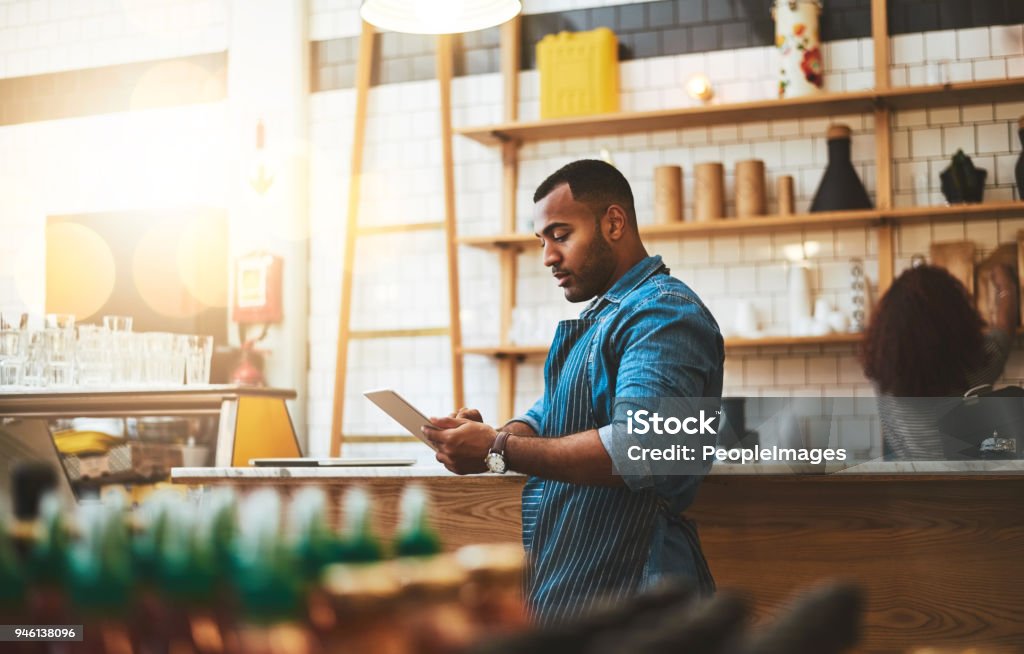 He's always making little tweaks to the business model Cropped shot of a handsome young man working on a tablet in his coffee shop Small Business Stock Photo