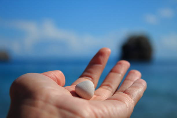 A close-up shot of a clenched hand holding some coarse sand that slowly falls to the ground, which is also covered with sand. The sand is not very fine, but consists of tiny pebbles. The image conveys a sense of time passing, loss or impermanence.