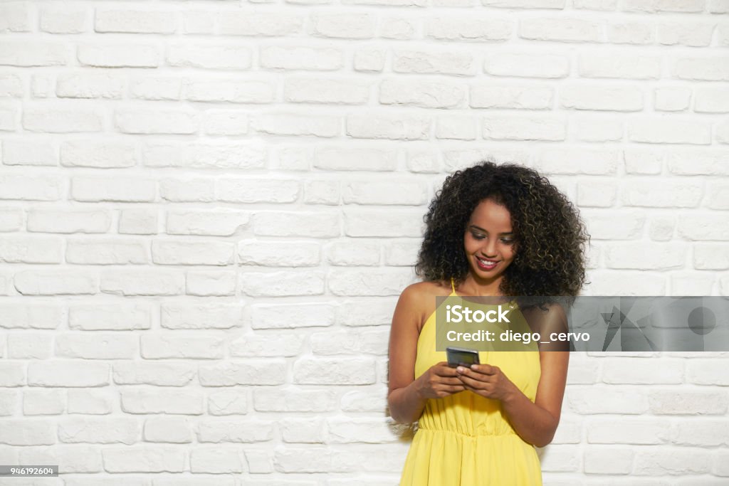 Facial Expressions Of Young Black Woman On Brick Wall Portrait of happy African American woman smiling against white brick wall and chatting on mobile phone. Mobile Phone Stock Photo