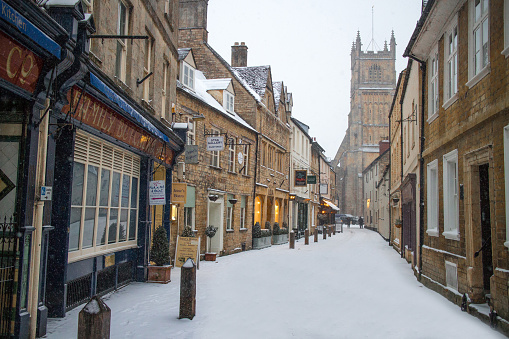 The small, quaint Blackjack Street in Cirencester, The Cotswolds with small shops and the parish church in the background, covered in snow