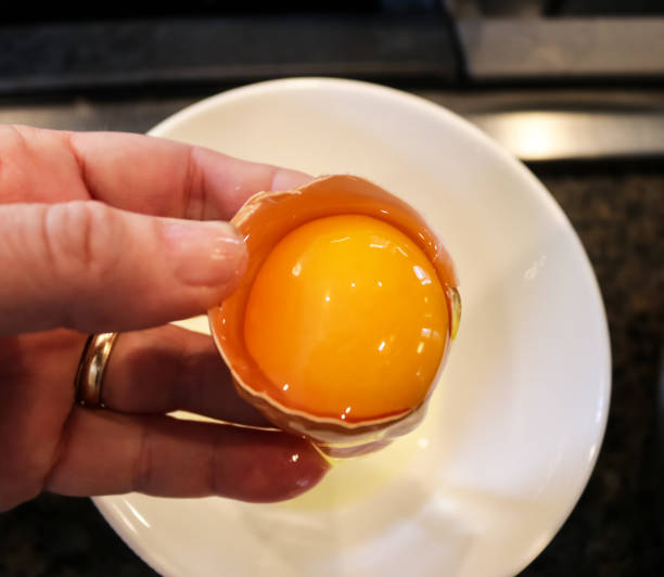 istock Separating an egg yolk from the egg white - hand holding half egg shell with yolk in it over white bowl containing the egg white - selective focus 946251628