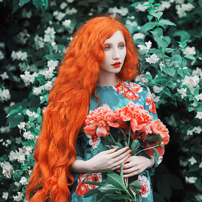 Woman with pale skin and long red hair in peony dress on background of a flower garden. Beautiful girl with red lips with a bouquet of peonies in hands. Beauty makeup. Redhead model. Sensual portrait