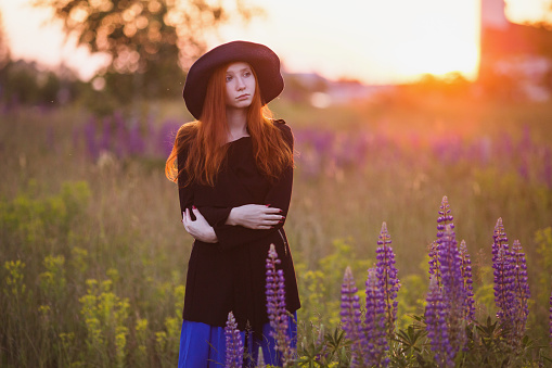 Redhead girl in a blue hat and black dress, with a backpack on her back in the backlit sunlight. The light from the setting sun. Life style