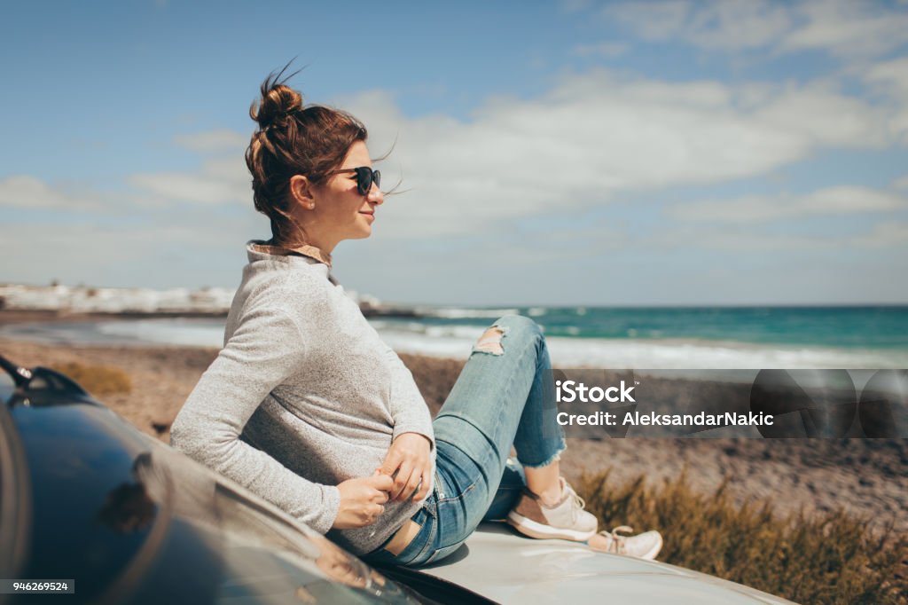 Young woman enjoys seaside view Photo of a young woman who enjoys seaside view, while on a road trip with her convertible car Car Stock Photo