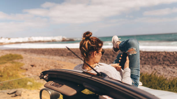Using a mobile phone during travel Photo of a young woman using a mobile phone while on a road trip with her convertible car by the seaside // wide photo dimensions hipster car stock pictures, royalty-free photos & images