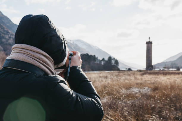 Man taking a photo of Glenfinnan Monument, Loch Shiel and Scottish landscape near Glenfinnan, Inverness-shire, Scotland. Man in winter clothes taking a photo of Glenfinnan Monument, Loch Shiel and Scottish landscape near Glenfinnan, Inverness-shire, Scotland, on a cold spring sunny day. glenfinnan monument stock pictures, royalty-free photos & images