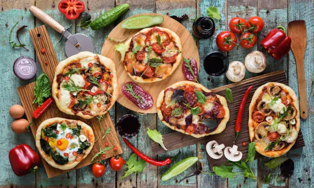 Flatlay of freshly prerared delicious Italian style pizzas. Five pizzas with eggs, salami, mushrooms and vegetables served with raw ingredients and red wine on shabby blue background overhead view