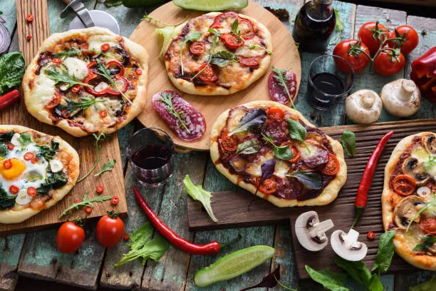 Top view of freshly prerared delicious pizzas. Italian style rustic pizzas with eggs, salami, mushrooms and vegetables served with raw ingredients and red wine on shabby blue background overhead view