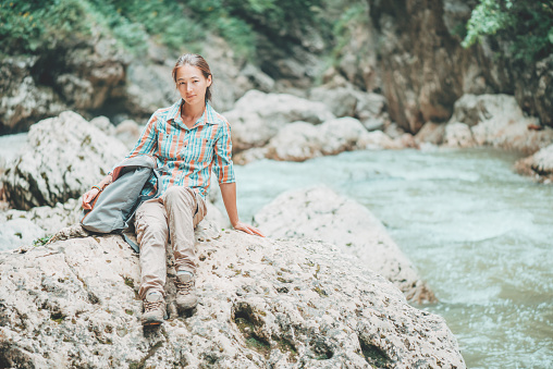 Backpacker young woman sitting on rocky stone on the river bank , looking at camera.