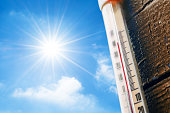 Thermometer with a high temperature reading on a scale, against a background of bright sun and a blue sky with clouds. The concept of hot, dangerous weather, global warming.