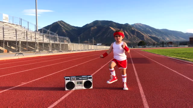 A young boy dressed in a headband and retro workout attire is warming up on a sports track before his workout. He is jumping, running in place and is excited for a healthy lifestyle. Video taken in Spanish Fork, Utah, USA.