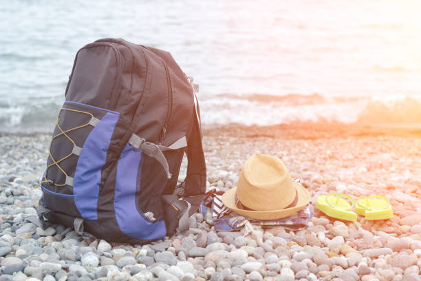 istock Backpack, hat and flip-flops on the pebble beach. Sea in the background 946365898