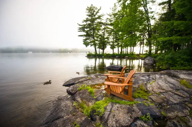 Adirondack chairs on a rock formation facing a calm lake. Ducks are in the water. The morning mist is covering part of the lake.