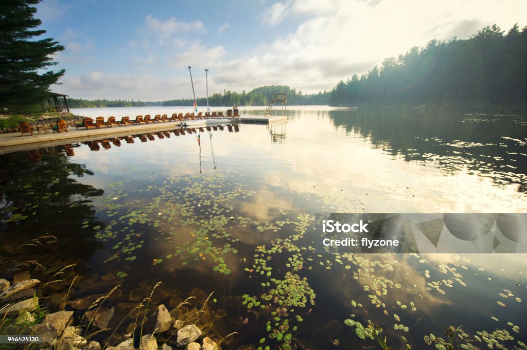 Adirondack and lounge chairs sitting on a dock on a lake Adirondack and lounge chairs sitting on a wooden dock facing a calm lake during a sunny summer day. Sailing boats are tied to the dock. Algonquin Provincial Park Stock Photo