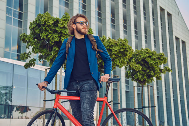 istock A handsome bearded man in stylish jeans and jacket and sunglasses with a backpack, standing with a bicycle on the street. 946458328