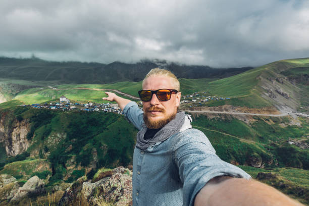 Global Tavel Concept. Young Traveler MAn With A Beard And Sunglasses Take A Selfie On A Background Of A Mountain Landscape stock photo