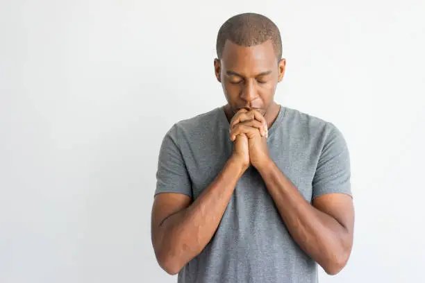 Calm spiritual handsome African guy praying with closed eyes. Serious peaceful young man with joining hands meditating. Belief concept
