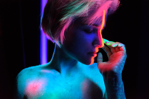 Young beautiful girl model with short haircut in the studio on a black background. Neon paints, makeup, body painting, colored hair coloring, creative color, poses in the light of soffits. Hands, face, blue, green, orange.