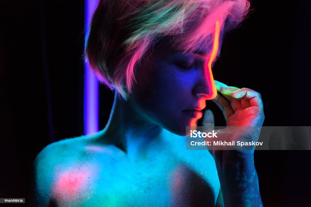 Model young beautiful girl portrait in studio with creative makeup, in neon ultraviolet lamp. Glows in the dark. Hair coloring with neon paints. Body art is blue, green, orange Young beautiful girl model with short haircut in the studio on a black background. Neon paints, makeup, body painting, colored hair coloring, creative color, poses in the light of soffits. Hands, face, blue, green, orange. Abstract Stock Photo