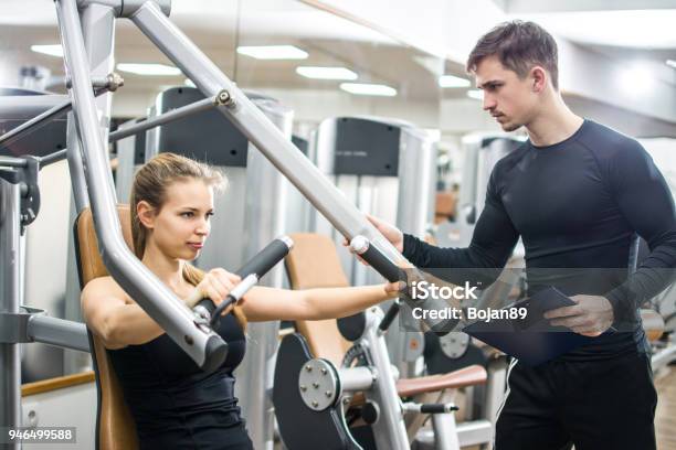 Sporty Girl Training On Exercise Machine With Support Of Her Personal Trainer In Gym Stock Photo - Download Image Now