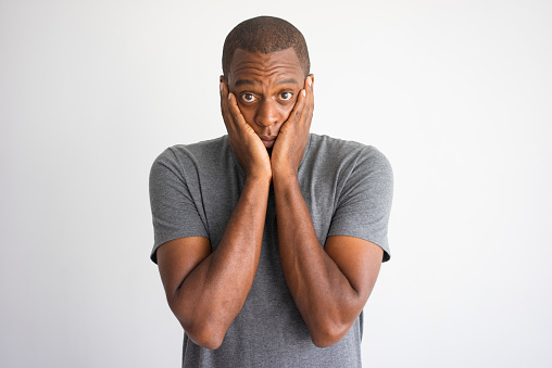 Portrait of shocked and puzzled African American. Stressed black man in casual wear surprised with bad news and covering cheeks with palms. Shock and bad news concept