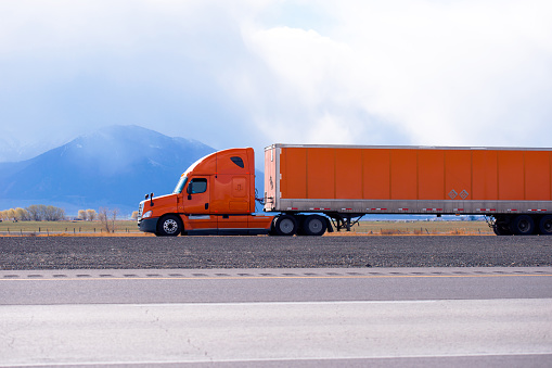Profile of orange bonnet big rig pro semi truck with long dry van orange semi trailer running on the flat road with blue mountain on background in Utah for long haul commercial cargo delivery trip