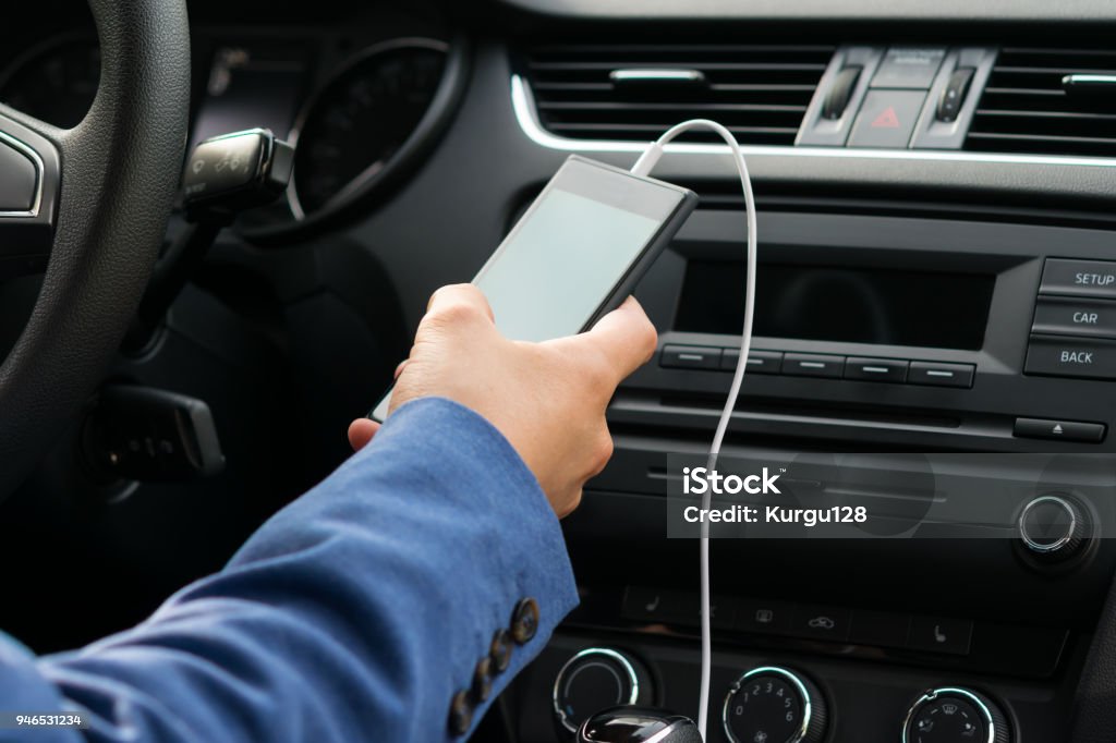 el conductor del vehículo, sostiene en su mano el teléfono conectado por un cable blanco, sistema de música del coche - Foto de stock de Cable libre de derechos