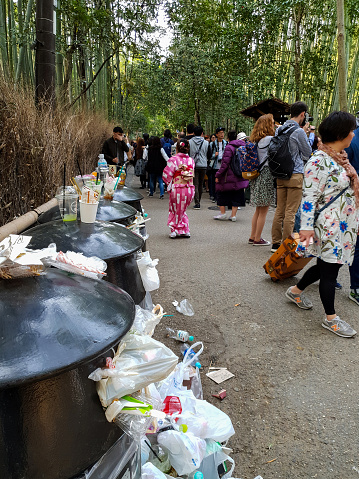 Japanese, Western and Asian tourists take pictures in the bamboo forest in Sagano, Kyoto during springtime, while in the foreground are overflowing recycle bins full of trash which has spilled around the bins due to the amount of tourists, Kyoto, Japan.