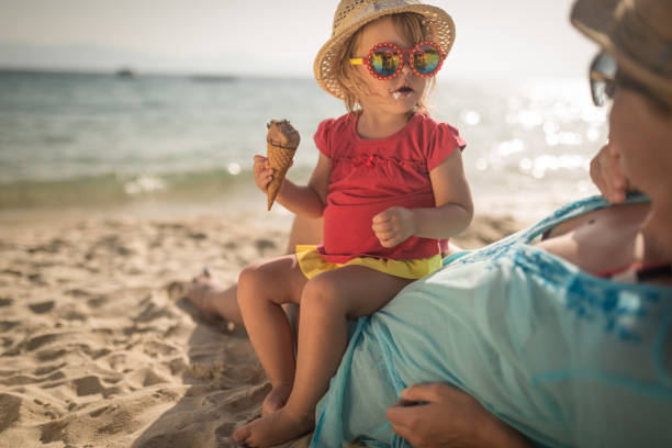 маленькая девочка сидения с матерью на песчаном пляже едят мороженое - fun mother sunglasses family стоковые фото и изображения