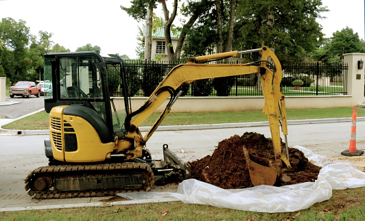 Yellow Backhoe with a pile of dirt on plastic parked on an upscale neighborhood Street with nice house a fence in the badkground